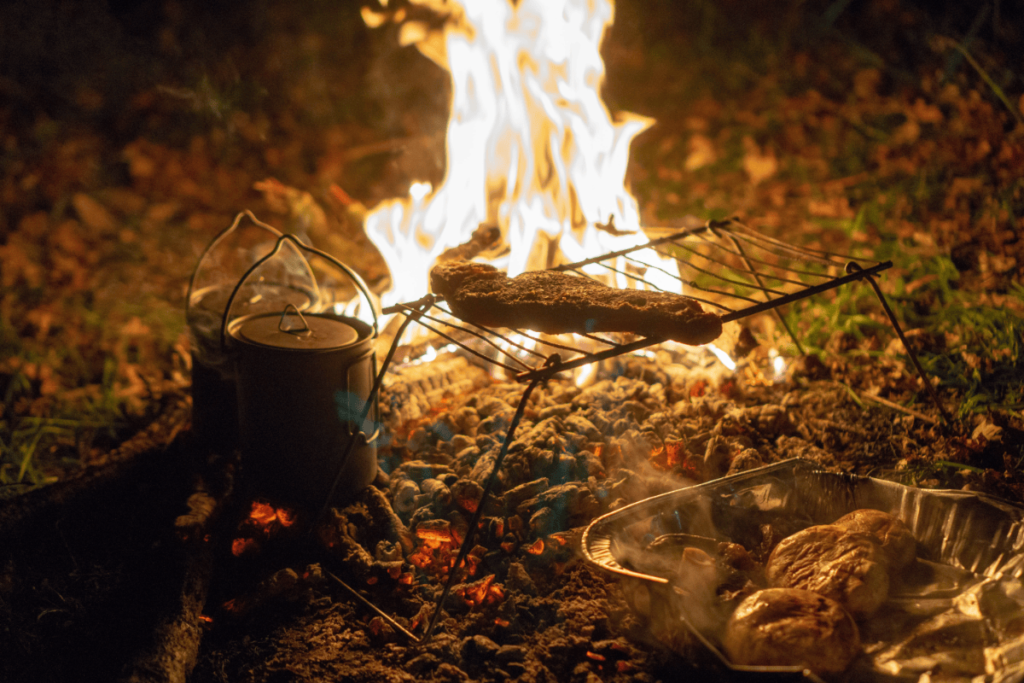 a bushcraft photo of a camp fire with water heating in a kettle and a steak on a light-weight foldable rack over hot coals. 